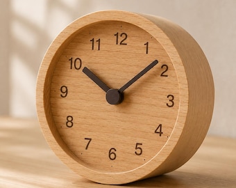 Könnte beinhalten: Eine runde analoge Uhr aus hellem Holz mit sichtbarer Maserung. Das Zifferblatt zeigt dunkelbraune Ziffern von eins bis zwölf sowie zwei dunkelbraune Zeiger, die die Zeit anzeigen. Das Design ist schlicht, kreisförmig und freistehend.