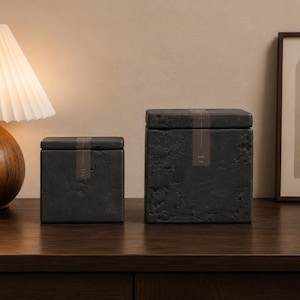 May include: Two square, dark gray stone containers with lids, each featuring a textured, porous surface and a vertical brown paper label centered on the front. The containers are of different heights, with the larger one positioned to the right.