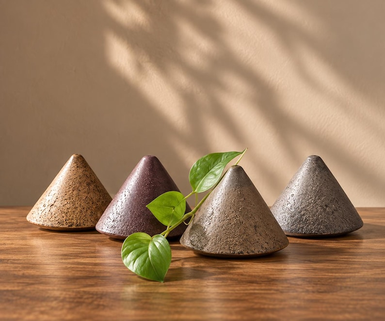 May include: Four cone-shaped ceramic objects with textured, speckled surfaces in shades of tan, dark purple, brown, and gray. A single green leafy vine rests across the front of the brown cone, which features a small opening at the top.