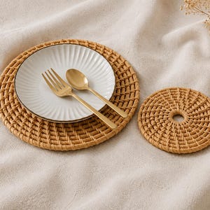 Puede incluir: Un mantel individual redondo de ratán trenzado con un plato de cerámica blanca de textura acanalada, un tenedor de metal dorado y una cuchara de metal dorado. Junto al mantel hay un posavasos redondo de ratán trenzado con una abertura central.