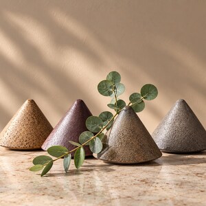 May include: Four cone-shaped ceramic objects with textured, speckled surfaces in shades of tan, dark purple, light brown, and gray. A single sprig of green eucalyptus leaves rests across the front of the light brown cone-shaped object.
