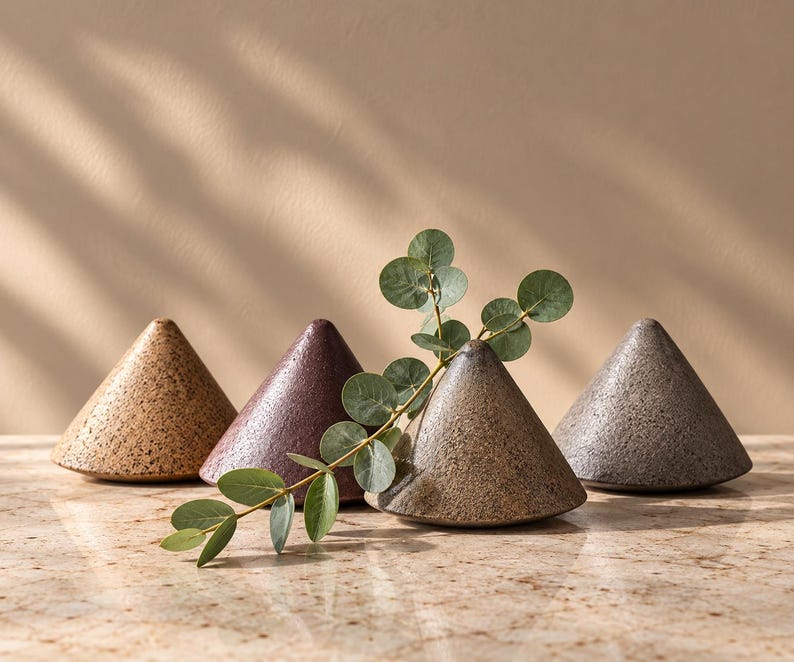 May include: Four cone-shaped ceramic objects with textured, speckled surfaces in shades of tan, dark purple, light brown, and gray. A single sprig of green eucalyptus leaves rests across the front of the light brown cone-shaped object.