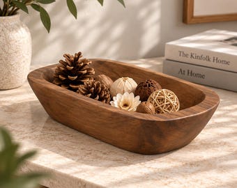 Könnte beinhalten: Eine ovale Holzschale mit glatter, dunkelbrauner Oberfläche, die eine Sammlung dekorativer Gegenstände enthält, darunter zwei Tannenzapfen, eine weiße Blume, eine geflochtene Rattankugel und mehrere kleine, runde, strukturierte braune botanische Kugeln.