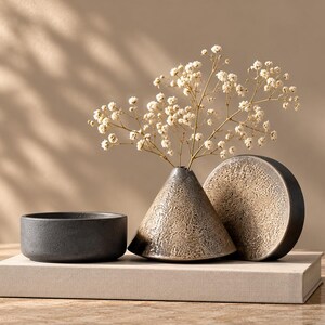May include: A small dark gray bowl, a conical vase with a textured beige and brown finish containing white baby's breath flowers, and a circular disc with a matching textured surface, all arranged together on a rectangular beige book.