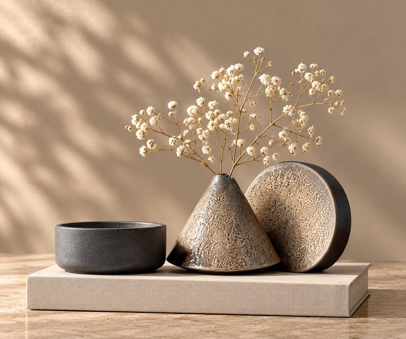 May include: A small dark gray bowl, a conical vase with a textured beige and brown finish containing white baby's breath flowers, and a circular disc with a matching textured surface, all arranged together on a rectangular beige book.