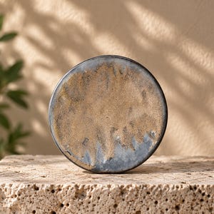 May include: A circular ceramic plate featuring a textured surface with a mottled blend of earthy tan, beige, and dark charcoal gray tones. The edges are defined by a dark, smooth rim, creating a distinct contrast against the rough, sandy center.