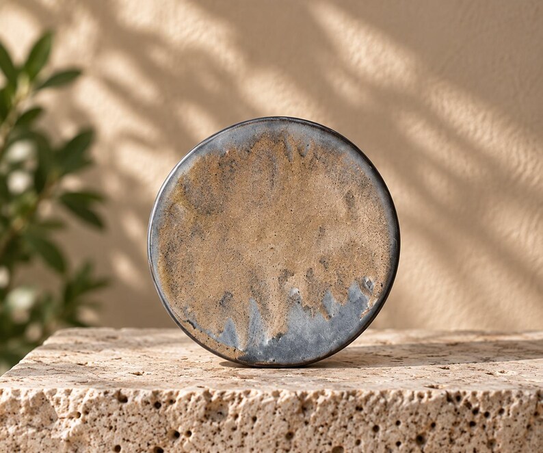 May include: A circular ceramic plate featuring a textured surface with a mottled blend of earthy tan, beige, and dark charcoal gray tones. The edges are defined by a dark, smooth rim, creating a distinct contrast against the rough, sandy center.