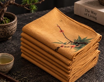 Könnte beinhalten: Ein Stapel senfgelber Stoffservietten mit einem aufgedruckten Motiv aus einem braunen Zweig mit grünen Blättern und kleinen rosa Blüten. Die Servietten sind rechteckig gefaltet und weisen eine texturierte Oberfläche auf dem Stoff auf.