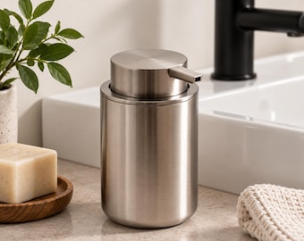 Könnte beinhalten: Ein zylindrischer Seifenspender aus gebürstetem Edelstahl mit flacher Oberseite und einem horizontalen, rechteckigen Auslauf. Der Spender hat eine glatte Metalloberfläche und ein schlichtes, funktionales Design für flüssige Seife.