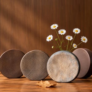 May include: Four circular, flat-faced objects with textured, earthy brown and grey surfaces arranged in a row. One object serves as a vase, holding a small bouquet of white daisies with yellow centers. A single dried leaf lies in the foreground.