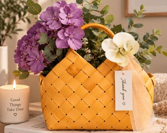 Könnte beinhalten: Ein gelber geflochtener Korb gefüllt mit violetten Hortensienblüten und grünen Eukalyptuszweigen. Eine weiße Orchidee und ein rechteckiges Schild mit dem Text "Thank you" und einem Herzsymbol sind mit einem pfirsichfarbenen Band befestigt.