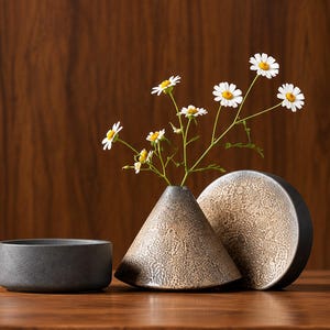 May include: A small, conical ceramic vase with a textured, speckled beige and brown finish, holding a sprig of white daisies with yellow centers. Beside it sits a matching circular, flat-topped ceramic object and a small, dark gray cylindrical bowl.