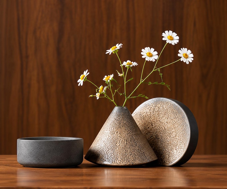 May include: A small, conical ceramic vase with a textured, speckled beige and brown finish, holding a sprig of white daisies with yellow centers. Beside it sits a matching circular, flat-topped ceramic object and a small, dark gray cylindrical bowl.