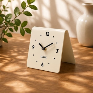 May include: A cream-colored, tent-shaped analog clock with a smooth, curved surface. The clock face features black numerals at 3, 6, 9, and 12, black dots for other hour markers, black hands, and the word CLOCK printed in black below the center.