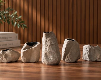 Könnte beinhalten: Ein Set aus fünf Keramikvasen mit einer strukturierten, hellbeigen Oberfläche, die an Naturstein erinnert. Jedes Gefäß hat eine unregelmäßige, organische Form mit einer dunklen, glatten Innenöffnung und variiert in Höhe und Breite.