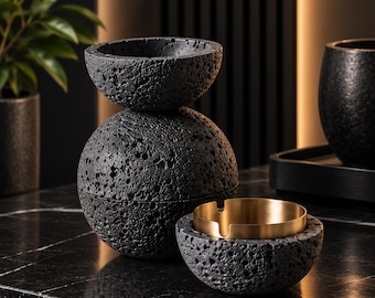 Könnte beinhalten: Ein Set aus dunkelgrauen, porösen Objekten mit vulkanischer Gesteinstextur, bestehend aus einem kugelförmigen Behälter mit Deckel, einer kleinen Schale und einer schalenförmigen Basis mit einem glatten, goldfarbenen Metalleinsatz mit zwei Kerben.