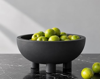 Könnte beinhalten: Eine matte, schwarze, runde Schale mit Füßen, gefüllt mit einer Ansammlung hellgrüner Limetten. Die Schale verfügt über ein glattes, tiefes Becken und wird von drei kurzen, zylindrischen Beinen gestützt, die eine stabile Basis für die Früchte bilden.