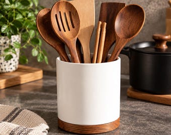 Könnte beinhalten: Ein zylindrischer Utensilienhalter aus weißer Keramik mit einem Boden aus Naturholz, der verschiedene Küchenhelfer aus Holz enthält, darunter einen Schlitzlöffel, einen Löffel, einen flachen Pfannenwender und ein Paar Essstäbchen.