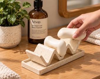 Könnte beinhalten: Eine beige gesprenkelte Seifenschale mit wellenförmigem, mehrstufigem Design, die zwei glatte, cremeweiße Seifenstücke hält. Der rechteckige Boden hat eine passende gesprenkelte Oberfläche und bietet eine stabile Basis.