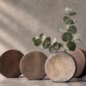 May include: Four circular, flat-faced vases with textured surfaces in shades of brown, copper, and gray. One vase contains a green eucalyptus branch with round leaves. The vases have a rounded, disc-like shape and a matte, earthy finish.