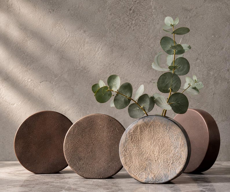 May include: Four circular, flat-faced vases with textured surfaces in shades of brown, copper, and gray. One vase contains a green eucalyptus branch with round leaves. The vases have a rounded, disc-like shape and a matte, earthy finish.