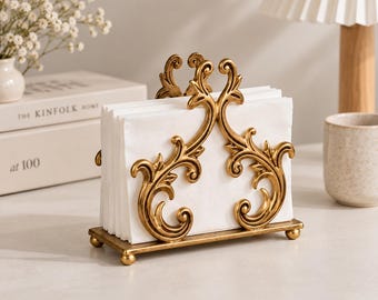 Könnte beinhalten: Ein goldfarbener Serviettenhalter aus Metall mit einem verzierten, symmetrischen Schnörkeldesign. Der Halter hat einen rechteckigen Sockel mit vier kleinen kugelförmigen Füßen und enthält einen Stapel weißer Papierservietten.
