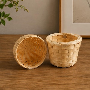 May include: Two small, round, light-colored woven baskets made from thin, flexible strips of wood. One basket is positioned upright, showing its open interior and woven base, while the other is placed on its side, revealing the exterior weave.