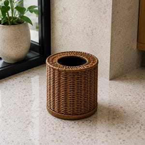 May include: A cylindrical tissue box cover featuring a woven brown rattan-style texture. The item has a circular opening at the top and a solid base, designed to hold a standard tissue box. The exterior displays a consistent, detailed weave pattern.