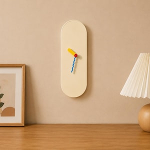 May include: A vertical, pill-shaped wall clock with a smooth, cream-colored face. The clock features a short, thick yellow hour hand and a longer, thin, blue wavy minute hand, both connected at the center by a small, circular red cap.