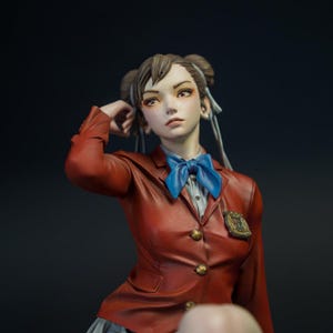 May include: A detailed figurine of a character with brown hair styled in two buns, wearing a red blazer with gold buttons, a blue bow tie, and a grey skirt. The character has a neutral expression and is posed with one hand near their head.