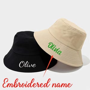 Könnte beinhalten: Ein schwarzer Fischerhut mit dem weiß gestickten Namen Olive und ein beigefarbener Fischerhut mit dem grün gestickten Namen Olivia. Ein orangefarbener Pfeil zeigt vom Text Embroidered name auf die grüne Stickerei auf dem beigen Hut.
