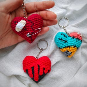 May include: Three heart-shaped crochet keychains. One red heart features a white bandage and a silver safety pin. Another red heart has a black dripping effect. The third heart is multicoloured with turquoise, orange, and pink sections.
