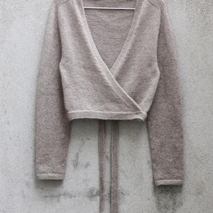 May include: A light taupe knit wrap top featuring long sleeves, a deep V-neckline, and a cropped length. The garment includes two long, thin knitted ties extending from the waist area, designed to wrap around the torso and secure at the front.
