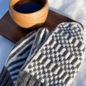 Könnte beinhalten: Ein Paar gestrickte Fäustlinge mit einem weiß-dunkelgrauen geometrischen Muster und gerippten Bündchen, platziert neben einer Holzschale mit dunkler Flüssigkeit und einem rechteckigen Holzbrett, alles auf einer weißen Schneefläche.