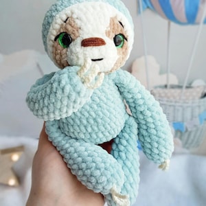 May include: A handmade crochet sloth plush toy crafted from soft, light blue and white yarn. The sloth features large, bright green plastic eyes, a brown stitched nose, and a small black mouth. It has long arms with white claws and a textured finish.