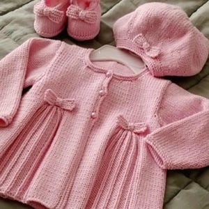 May include: A light pink knitted set consisting of a long-sleeved cardigan with three buttons and two decorative bows, a matching beret with a small bow, and a pair of baby booties featuring bows on the front, all crafted in a textured knit.