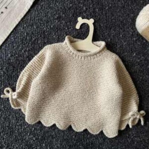 May include: A cream-coloured knit jumper featuring a ribbed crew neckline, long sleeves with tied cuffs, and a scalloped hemline. The garment is displayed on a light-coloured hanger, with a ball of yarn labelled Soft Tweed positioned nearby.