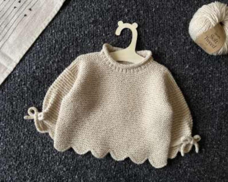 May include: A cream-coloured knit jumper featuring a ribbed crew neckline, long sleeves with tied cuffs, and a scalloped hemline. The garment is displayed on a light-coloured hanger, with a ball of yarn labelled Soft Tweed positioned nearby.