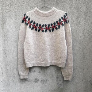 Può includere: Un maglione in maglia beige a maniche lunghe con scollo rotondo e un motivo decorativo sullo sprone. Lo sprone presenta un design ripetitivo di foglie verde scuro e piccole bacche rosse, realizzato con un dettaglio a maglia testurizzata.