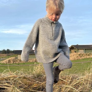May include: A light gray long-sleeve knit sweater featuring a high ribbed collar with a short front zipper closure, paired with black and white horizontally striped leggings and dark gray ankle boots, worn in an outdoor setting.