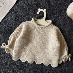 May include: A cream-colored knit sweater featuring a ribbed crew neckline, long sleeves with tied cuffs, and a scalloped hemline. The garment is displayed on a light-colored hanger, with a ball of yarn labeled Soft Tweed positioned nearby.
