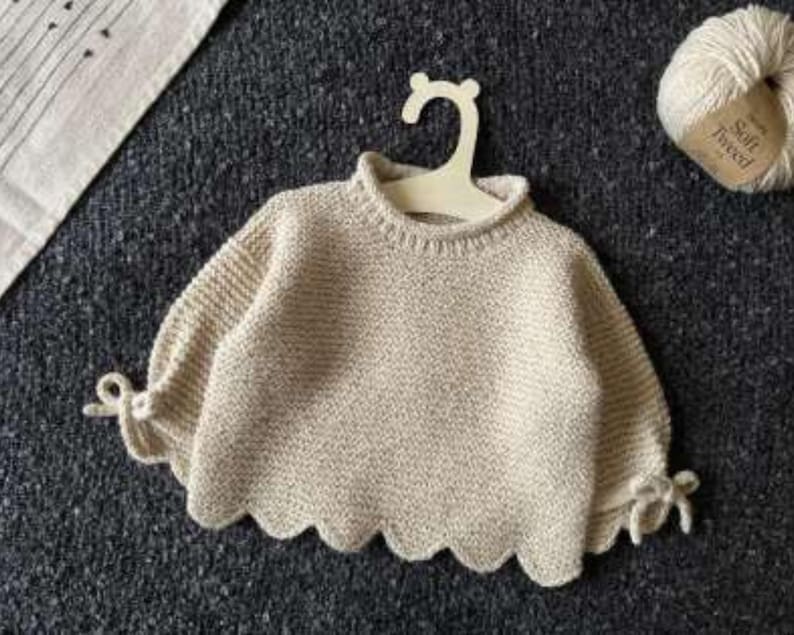 May include: A cream-coloured knit jumper featuring a ribbed crew neckline, long sleeves with tied cuffs, and a scalloped hemline. The garment is displayed on a light-coloured hanger, with a ball of yarn labelled Soft Tweed positioned nearby.