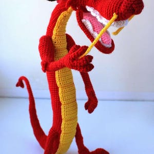 May include: A crocheted dragon figurine featuring a vibrant red body with a long yellow underbelly, a wide open mouth with white teeth, a pink tongue, and black eyes. The creature has long yellow whiskers, a thin tail, and small horns.