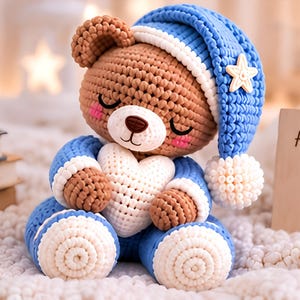 Könnte beinhalten: Ein gehäkelter brauner Teddybär mit geschlossenen Augen und rosa Wangen, der eine blaue Schlafmütze mit weißem Bommel und einem gelben Stern trägt. Der Bär hält ein weißes gehäkeltes Herz und trägt einen blauen Schlafanzug mit weißen Bündchen.
