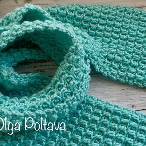 May include: A turquoise crochet scarf with a textured stitch pattern. The scarf is folded and draped, showcasing its length and the intricate details of the handmade design. The scarf is made of soft yarn.