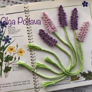 May include: Crocheted lavender flower bookmarks with green stems and tassels rest on an open book with botanical illustrations. The text "(c) Olga Poltava" is visible. The bookmarks are purple and pink.