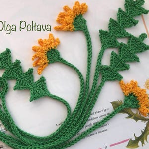 May include: A handmade crochet bookmark featuring a dandelion design. The bookmark has green stems and leaves, with yellow and orange flower heads. The bookmark is placed on an open book with text and illustrations.