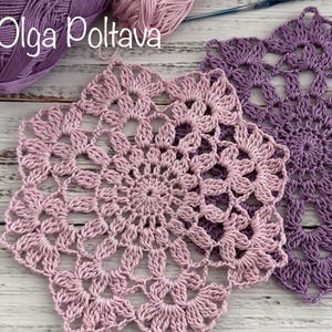 May include: Two handmade crochet doilies in shades of purple and pink, with a floral design. The doilies are placed on a light-colored wooden surface. A ball of purple yarn is visible in the background.