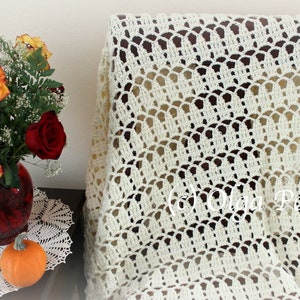 May include: A white crocheted throw blanket with a delicate, lacy pattern. The blanket is draped over a wooden chair and has a scalloped edge.