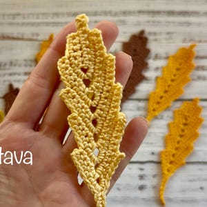 May include: A close-up of a hand holding a yellow crocheted leaf, with other crocheted leaves in shades of yellow and brown. The leaves are on a light-coloured wooden surface. The image includes the text "(c) Olga Poltava".